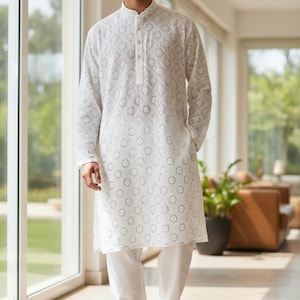 May include: A white, long-sleeved tunic and matching pants set. The tunic features a detailed, circular pattern and a mandarin collar. The outfit is paired with white sneakers. The person is standing in a well-lit room with large windows.