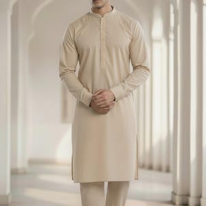 Puede incluir: Un conjunto de kurta pijama beige. La kurta de manga larga tiene cuello mandarín y abotonadura. Los pantalones a juego son de corte recto. El atuendo se usa con sandalias marrones. El fondo es un pasillo blanco con puertas arqueadas.