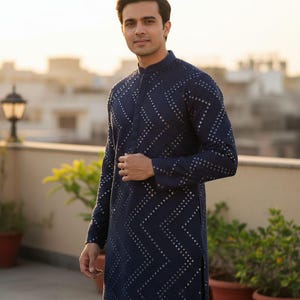 May include: Navy blue long-sleeved tunic with a mandarin collar and a geometric pattern of small, silver-colored embellishments. The tunic is knee-length and has a side slit. The person is standing outdoors.