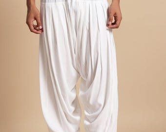 May include: White, loose-fitting pants with a drawstring waist and gathered fabric. The pants have a relaxed fit and are paired with black shoes. The text "CAFTAN GLAMOUR" is displayed in a white box at the bottom.