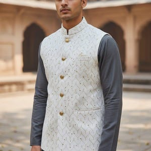 May include: A cream-colored, patterned vest with gold buttons and a mandarin collar. The vest is worn over a gray long-sleeved shirt and matching pants. The outfit is suitable for formal occasions.