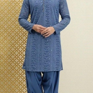 May include: A blue tunic and salwar suit with intricate paisley patterns. The long-sleeved tunic has a mandarin collar and button details. The matching salwar pants are loose-fitting. The outfit is worn with brown leather sandals.