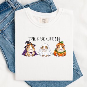May include: A white t-shirt featuring a graphic of three guinea pigs in Halloween costumes with the text TRICK OR WHEEK! above them. One guinea pig wears a witch hat and purple cape, one is under a white ghost sheet, and one is inside a pumpkin.