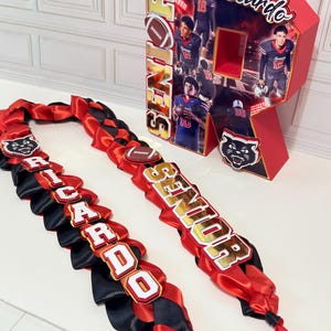 May include: A red, black, and gold ribbon lei featuring the name RICARDO and the word SENIOR, paired with a large, three-dimensional letter R display decorated with football-themed photos, the name Ricardo, and a black panther mascot logo.