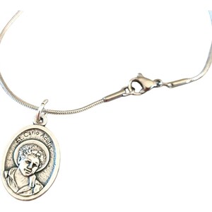 May include: A silver chain necklace with a silver pendant featuring a portrait of St. Carlo Acutis. The pendant is oval shaped and has the words "St. Carlo Acutis" inscribed around the edge.