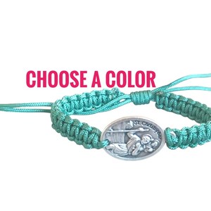 Könnte beinhalten: Ein türkisfarbenes Makramee-Armband mit einem ovalen Silberanhänger mit St. Christopher. Das Armband hat verstellbare Schiebeknoten und den Text "CHOOSE A COLOR" in Pink.