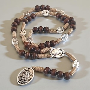 May include: A rosary featuring dark brown wooden beads and silver-coloured metal accents. The beads are strung on a beige cord. The rosary includes oval pendants with religious imagery, including one with the text "ST. MICHAEL".