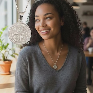 Puede incluir: Un collar con colgante plateado con un medallón redondo y detallado. El collar lo lleva una persona con un suéter gris con cuello en V. El medallón tiene un diseño intrincado y está suspendido de una delicada cadena. El fondo está borroso.