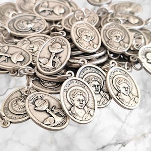 Może przedstawiać: Zbliżenie na stos srebrnych, owalnych medali z wizerunkami kielicha i świętego. Medale mają napis "St. Carlo Acutis".