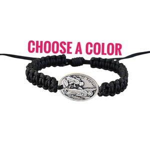 May include: Black braided bracelet with a silver oval charm featuring an image of Saint Raphael. The charm has the text "St. Raphael" inscribed on it.