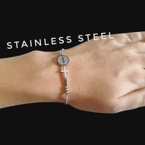 May include: Stainless steel bracelet featuring a round medallion and a cross pendant. The bracelet is silver-colored and is displayed on a person's hand against a black background. The word "STAINLESS STEEL" is written in white.