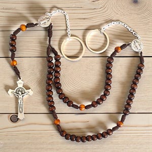 May include: A wooden rosary with dark brown beads and orange accent beads. The rosary features a silver crucifix and a silver medallion. The rosary is displayed on a light wood surface.
