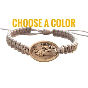 Puede incluir: Un brazalete trenzado beige con un dije ovalado dorado que representa a San Francisco de Asís. El dije tiene las palabras "St. Francis" inscritas. El brazalete tiene un cierre de nudo deslizante.
