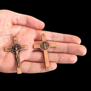 Puede incluir: Dos cruces de color bronce. Una cruz presenta una representación de Jesús en la cruz con las letras "INRI" arriba. La otra cruz tiene un emblema circular con una figura. Ambas cruces tienen un lazo en la parte superior para colgar.