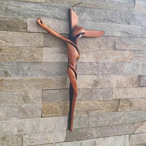 Puede incluir: Un crucifijo de madera con un diseño único y fluido. La cruz es de color marrón cálido y está montada en una pared de piedra texturizada. Los brazos de la cruz se extienden hacia afuera, y la figura tiene una forma estilizada y abstracta.