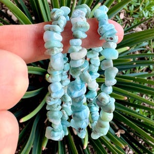 May include: Several strands of light blue and white gemstone chip bracelets. The bracelets are made of irregular, polished stones, strung together. The background features green foliage.