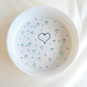 Puede incluir: Cuenco de cerámica blanco con un diseño dibujado a mano de corazones y puntos en verde azulado y azul. Un gran corazón está centrado en el cuenco, rodeado de corazones más pequeños y puntos azules. El cuenco tiene una forma simple y redondeada.