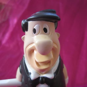 Può includere: Una statuetta di plastica di un personaggio dei cartoni animati che indossa un abito nero e un papillon. Il personaggio ha un cappello nero, capelli castani e un naso grande e da cartone animato.