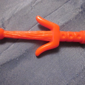 May include: Orange plastic sai, a traditional weapon used in martial arts. The sai has a rounded tip and a textured handle.