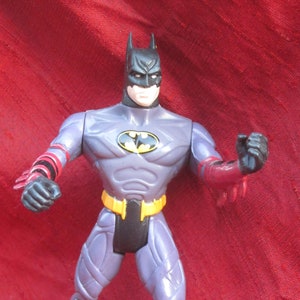 May include: A Batman action figure in a dynamic pose. The figure is mainly grey and purple, with black accents on the boots, gloves, and cowl. The bat symbol is on the chest, with a yellow belt and red details on the arms and legs.