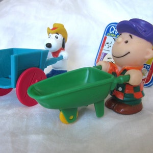 Könnte beinhalten: Ein grüner Spielzeug-Bollerwagen mit einem gelben Rad, ein blauer Spielzeug-Bollerwagen mit einem roten Rad und eine Plastikfigur von Charlie Brown in einem karierten Hemd und einer lila Mütze.