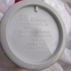 May include: White ceramic mug with a blue, yellow, and red design. The bottom of the mug has the text "Eландия Santa Clarita CA 91355 Not for microwave or dish washer use Taiwan Swivel Mug U.S.A. Patent No. 5400907"