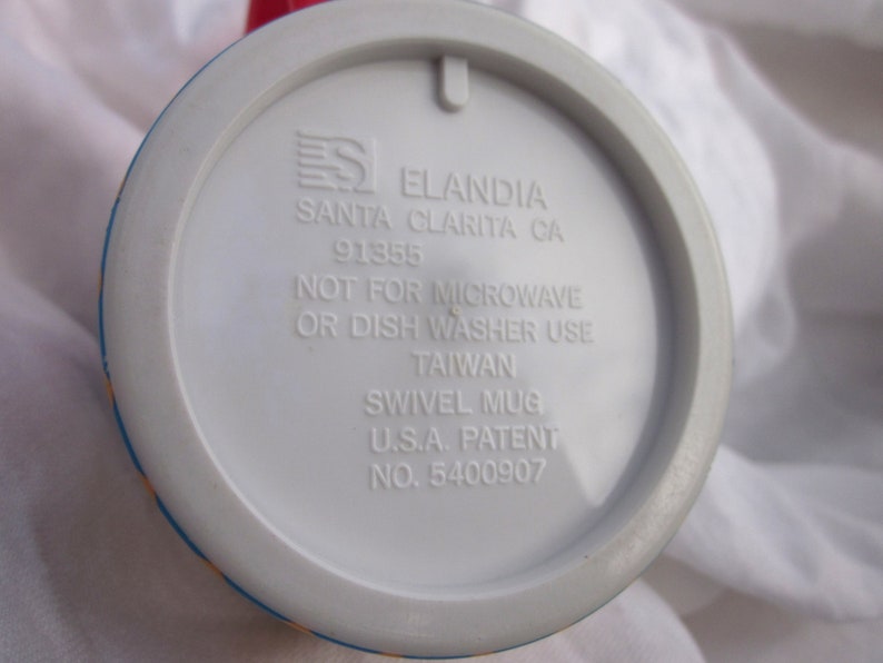 May include: White ceramic mug with a blue, yellow, and red design. The bottom of the mug has the text "Eландия Santa Clarita CA 91355 Not for microwave or dish washer use Taiwan Swivel Mug U.S.A. Patent No. 5400907"