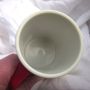 May include: A white ceramic mug with a red handle. The mug is empty and the inside is visible.