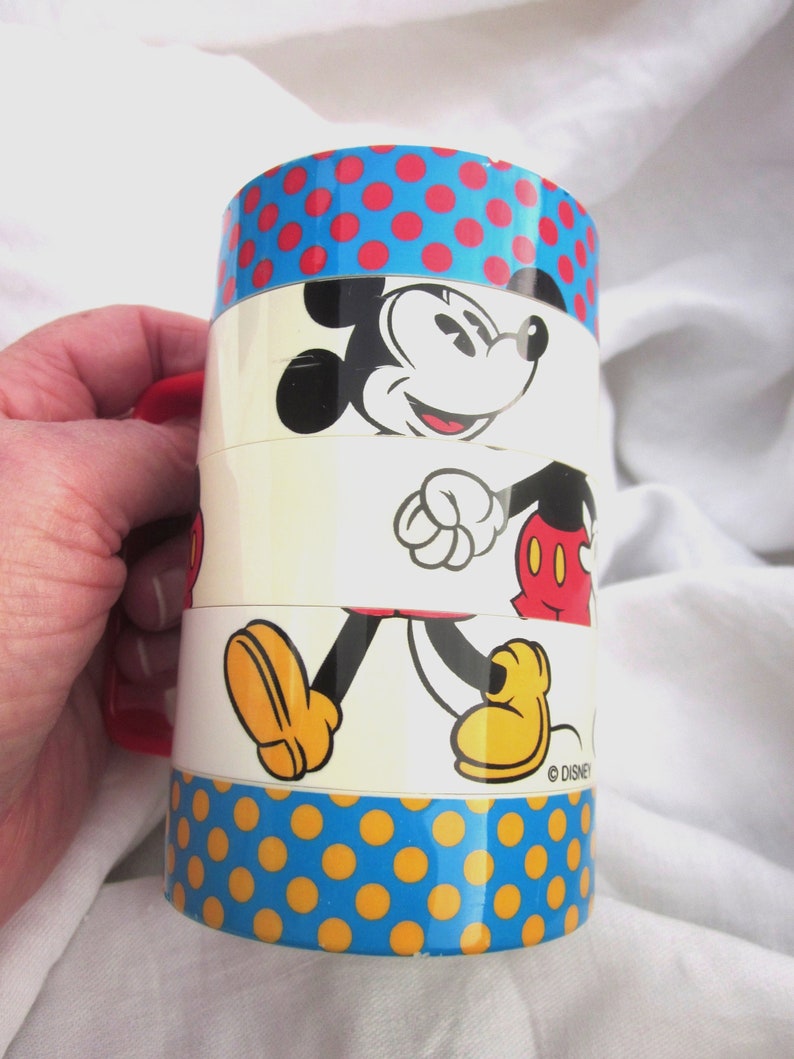 May include: A vintage Mickey Mouse stacking cup with a blue and yellow polka dot pattern. The cup features a red handle and a white background with a cartoon image of Mickey Mouse walking. The bottom of the cup has the text "&copy; DISNEY".