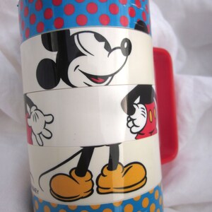 May include: A red and blue stacking cup with a Mickey Mouse design. The cup features a red polka dot pattern, a white section with Mickey Mouse's face, a white section with Mickey Mouse's legs, and a blue section with yellow polka dots. The cup has a red handle.