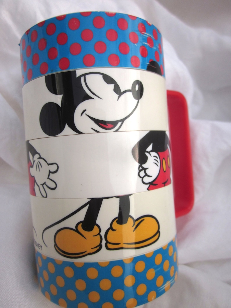 May include: A red and blue stacking cup with a Mickey Mouse design. The cup features a red polka dot pattern, a white section with Mickey Mouse's face, a white section with Mickey Mouse's legs, and a blue section with yellow polka dots. The cup has a red handle.