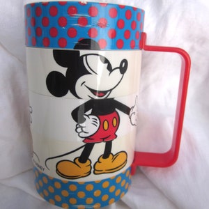 May include: A vintage Mickey Mouse mug with a red handle and a blue and red polka dot pattern. The mug features a cartoon image of Mickey Mouse in a red and yellow outfit. The bottom of the mug has a yellow and blue polka dot pattern.