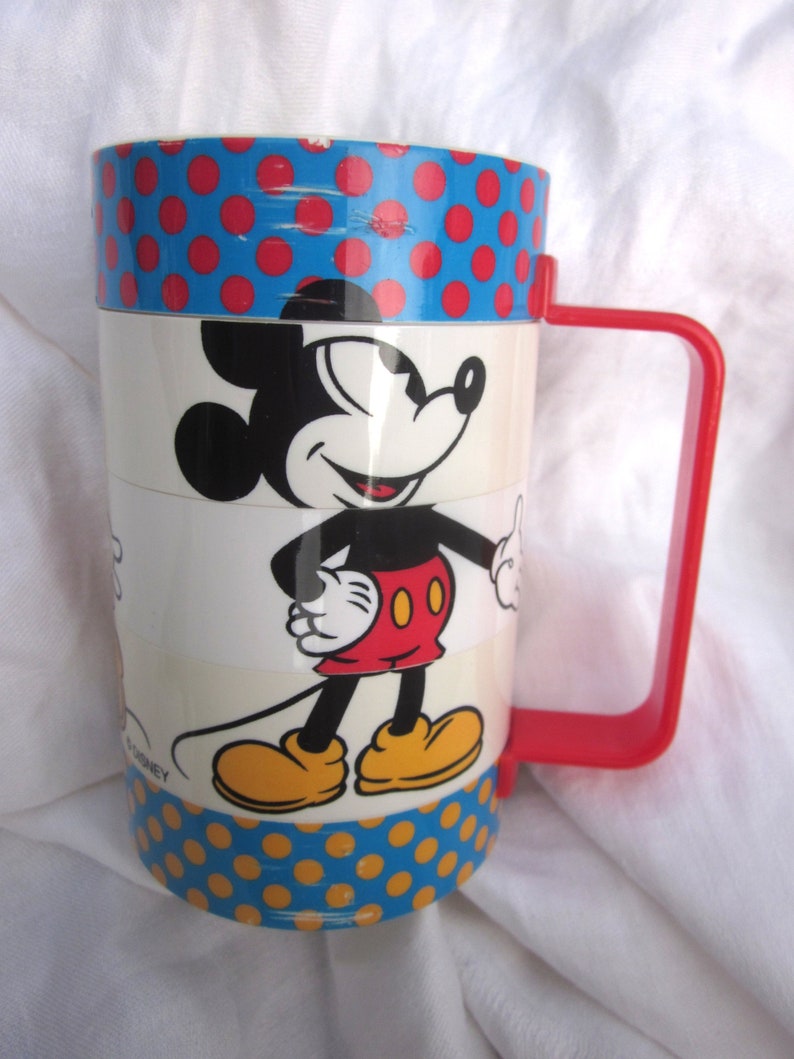 May include: A vintage Mickey Mouse mug with a red handle and a blue and red polka dot pattern. The mug features a cartoon image of Mickey Mouse in a red and yellow outfit. The bottom of the mug has a yellow and blue polka dot pattern.