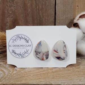 May include: A pair of handmade ceramic earrings with a white base and brown, blue, and gray marbling. The earrings are shaped like teardrops and are displayed on a white card with the text "JC Designs Clay Handmade Jewelry".