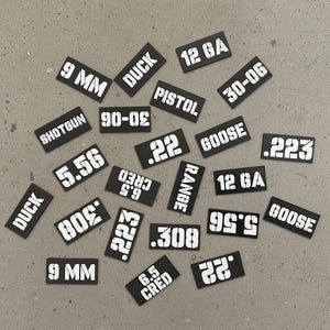 Könnte beinhalten: Eine Sammlung rechteckiger schwarzer Etiketten mit weißem Schablonentext, darunter 9 MM, DUCK, 12 GA, PISTOL, 30-06, SHOTGUN, 5.56, .22, GOOSE, .223, 6.5 CRED, RANGE, .308 und 6.5 CRED. Diese kleinen Marker haben eine strukturierte Oberfläche.