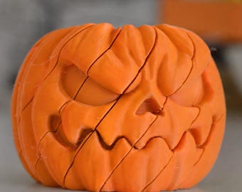 May include: A bright orange pumpkin-shaped object featuring a carved, menacing facial expression with deep, angular grooves and segmented lines across its surface, creating a puzzle-like appearance with a hollow top opening.