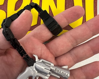 May include: A silver-colored revolver pendant with the word "Sparks" engraved on the barrel, connected to a black chain with a black clasp. The pendant is held in a hand.