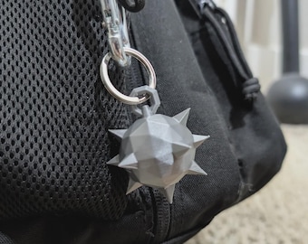 May include: A gray, multi-faceted, spiked mace-shaped keychain charm attached to a silver-colored metal split ring and a carabiner clip. The geometric, spherical object features several sharp, pointed protrusions extending from its surface.