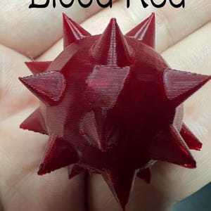 May include: A 3D printed, blood red, spherical object featuring multiple sharp, pointed spikes protruding from its surface. The text "Blood Red" is printed in black at the top of the image, clearly labelling the colour of the spiked sphere.