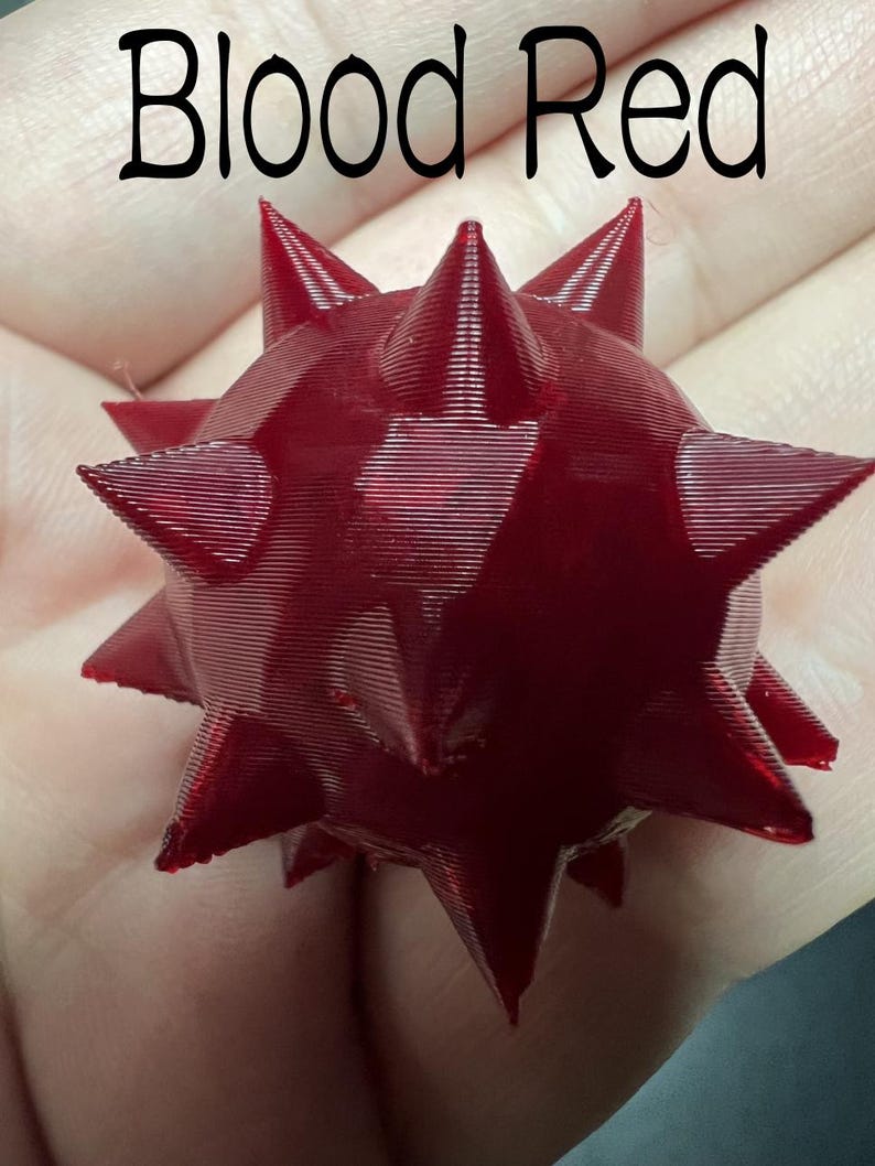 May include: A 3D printed, blood red, spherical object featuring multiple sharp, pointed spikes protruding from its surface. The text "Blood Red" is printed in black at the top of the image, clearly labelling the colour of the spiked sphere.