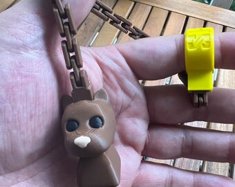 May include: A brown, 3D-printed bear pendant with a chain and a yellow ring. The bear has black eyes and a white nose. The chain and ring are also brown and yellow, respectively. The pendant is held in a hand.