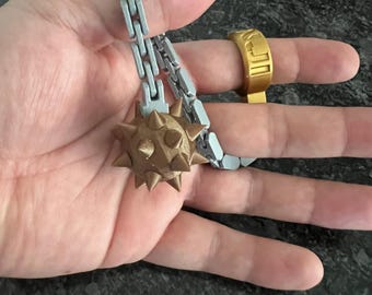 May include: A gold-colored ring and a silver chain with a brown spiked ball are held in a hand. The ring has a rectangular design. The ball has multiple spikes.