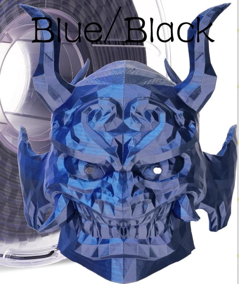 May include: A 3D printed mask with a geometric, faceted surface in a metallic blue and black finish. The mask features two prominent curved horns, a wide grin with visible teeth, and eye openings. The text Blue/Black is printed above.