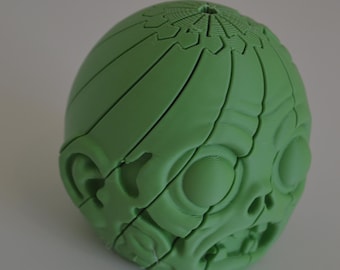 May include: A light green, spherical, 3D-printed puzzle object featuring a stylized face with prominent circular eyes, a small nose, and an open mouth. The surface is composed of interlocking segments that form a rounded, textured head shape.