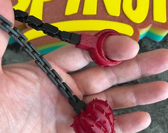 May include: A red spiky ball attached to a black chain and a red finger ring. The object is positioned in front of a yellow sign with the text SPiNJi. The red components feature a textured surface, while the chain consists of small black links.
