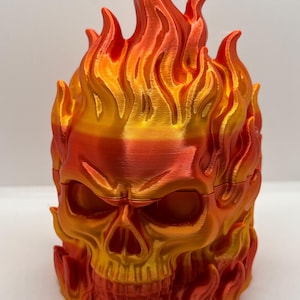Puede incluir: Una figura de calavera impresa en 3D con un degradado vibrante de colores naranja, rojo y amarillo metálicos. La calavera está rodeada de llamas esculpidas que se elevan hacia arriba, cubriendo la parte superior y los lados con textura.