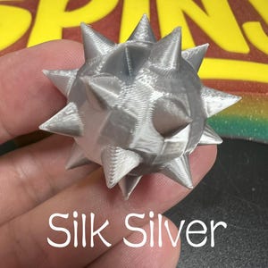 May include: A 3D printed silver sphere with multiple pointed spikes protruding from its surface. The object has a metallic sheen and a visible layered texture. The text Silk Silver is printed below the object, and the word SPINY is visible above.