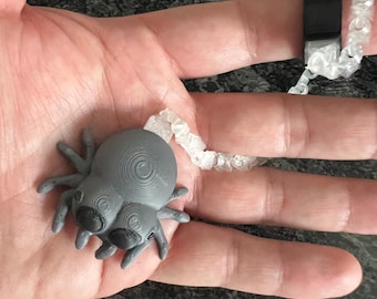 May include: A gray 3D-printed spider figurine with large black eyes, attached to a clear, textured, flexible chain that connects to a black ring band. The spider features visible concentric circular layer lines on its body and head.