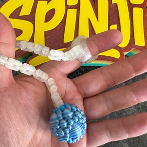 May include: A clear, plastic finger ring and beaded necklace with a blue and white striped orb. The word "SPINJI" is in yellow letters on a brown background.