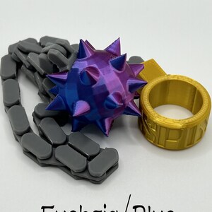 May include: A 3D-printed flail toy featuring a purple and blue spiked ball attached to a grey articulated chain and a gold ring. The text "Fuchsia/Blue" is printed at the bottom of the image. The object is composed of a spiked sphere, a flexible chain, and a circular ring component.